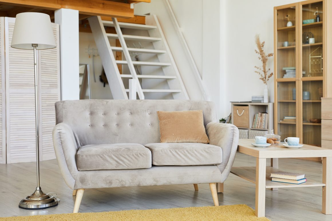 Canapé gris clair avec coussin beige, lampe sur pied argentée, table en bois clair avec tasses, livres et décor minimaliste.