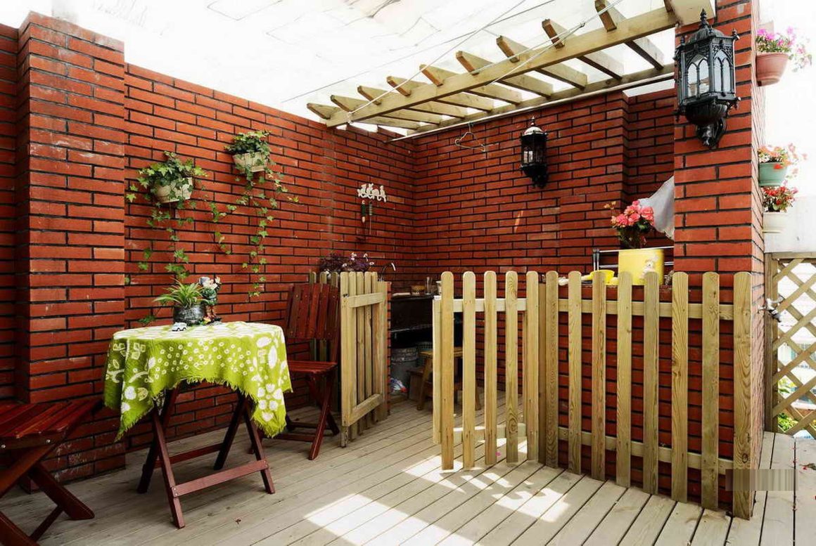 Petite terrasse avec mur en briques rouges, plantes et coin repas incluant table et chaises pliantes.