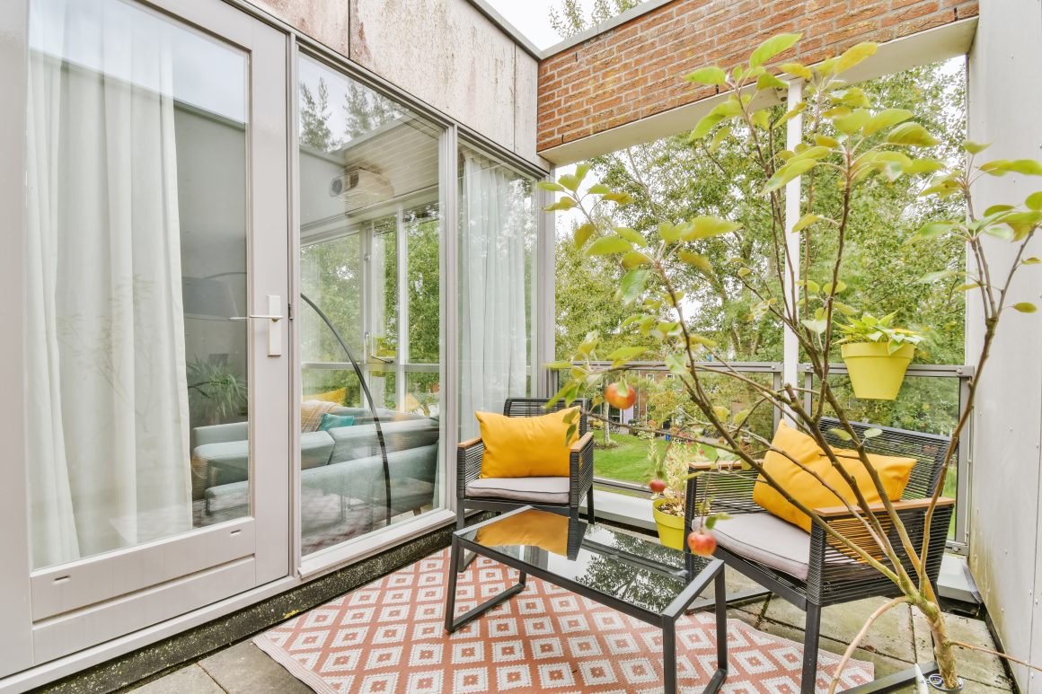 Aménagement petite terrasse avec chaises en rotin, coussins jaunes, table en verre, tapis à motifs, et plantes vertes.