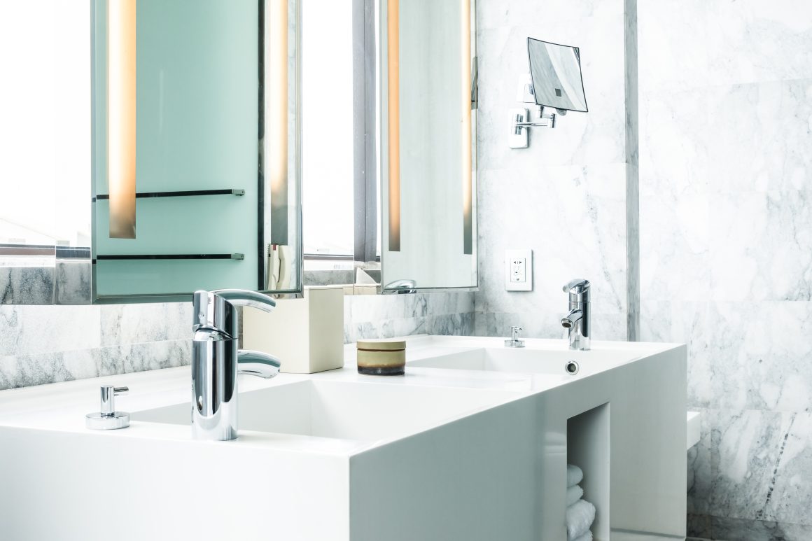 Salle de bain moderne avec double lavabo blanc, robinets chromés, miroir et murs en marbre.