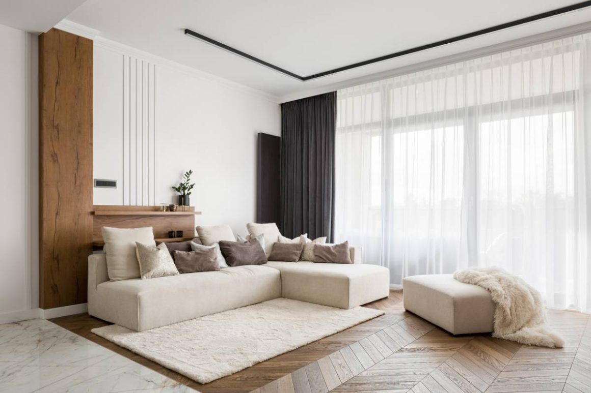 Canapé de salon beige, tapis blanc, fauteuil et grande fenêtre avec rideaux noirs et transparents.