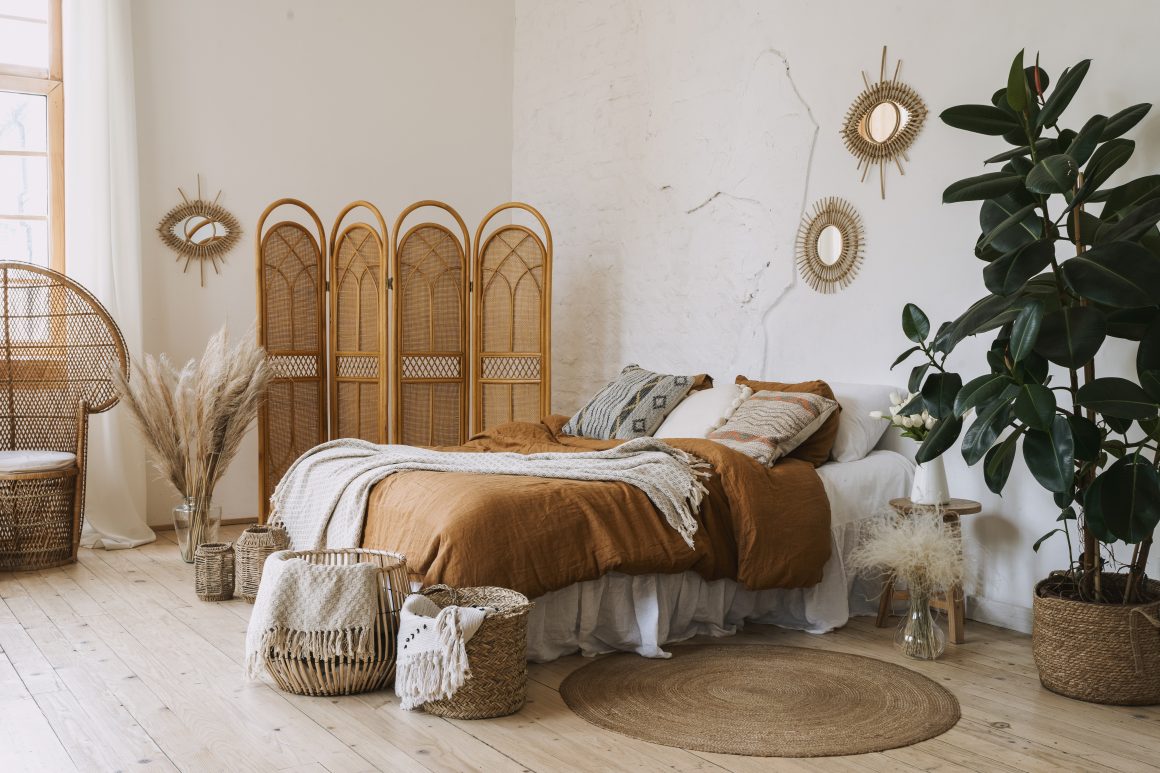 Chambre chaleureuse avec décoration bohème, meubles en osier, literie marron, coussins, plantes, tapis rond et mur blanc décoré.