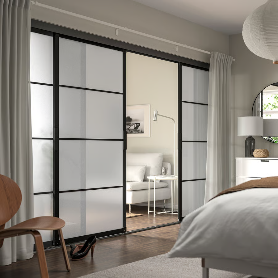 Chambre moderne avec portes coulissantes en verre, parquet foncé, fauteuil en bois et rideaux blancs.