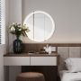 Un coin chambre moderne avec coiffeuse en bois, miroir rond lumineux, tabouret marron, fleurs blanches, et coussin gris clair.