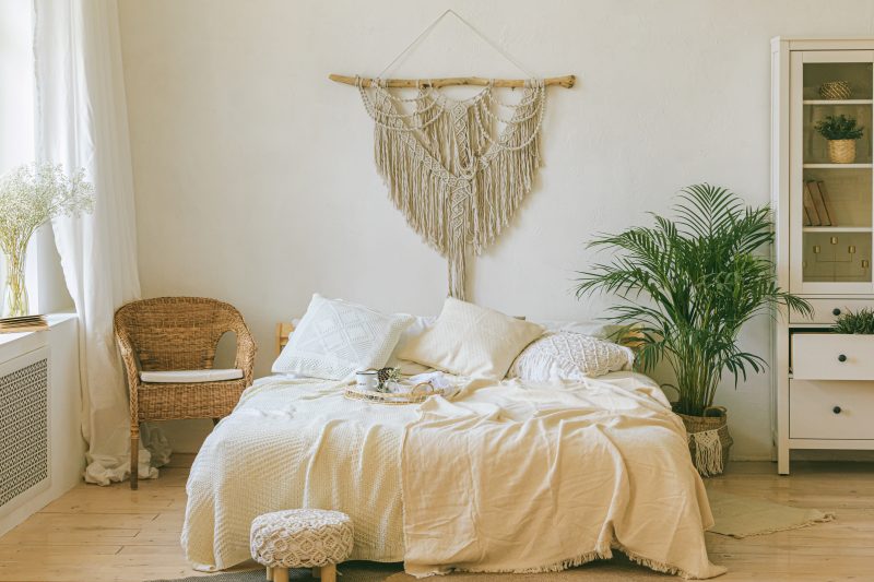 Chambre style bohème avec murs clairs, literie crème, fauteuil en osier, plante verte et tapis beige.