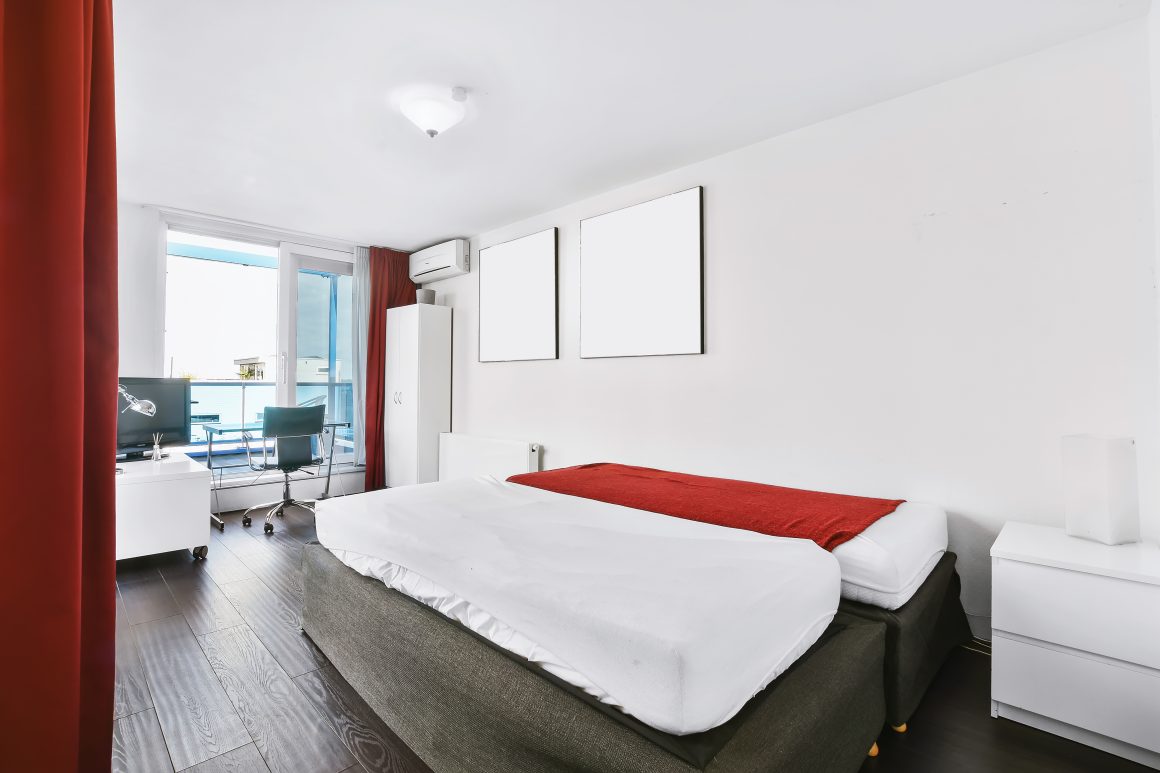 Studio incluant lit, rideau rouge, bureau, chaise avec parquet bois foncé et murs blancs.