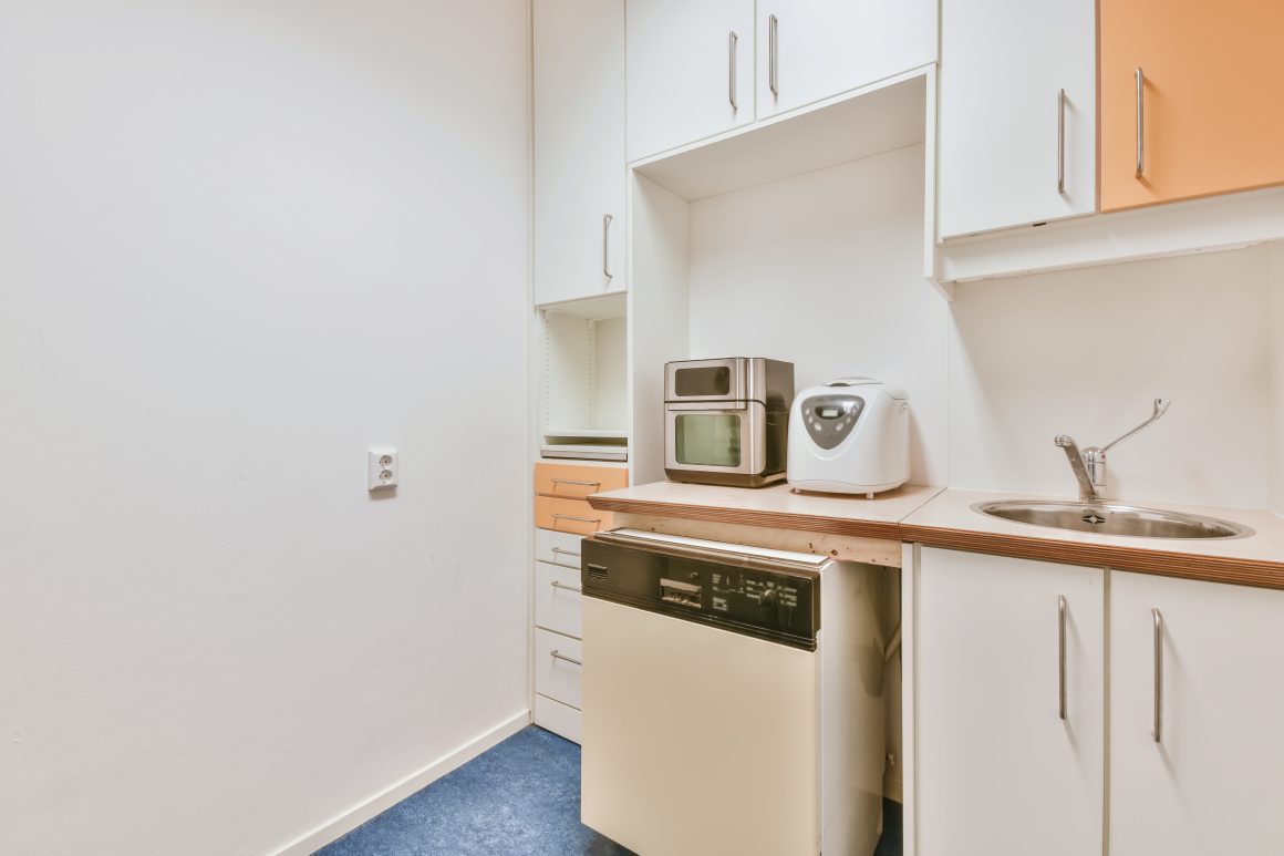 Petite kitchenette avec comptoir, micro-ondes, évier, armoires blanches, sol en moquette bleue et murs blancs.