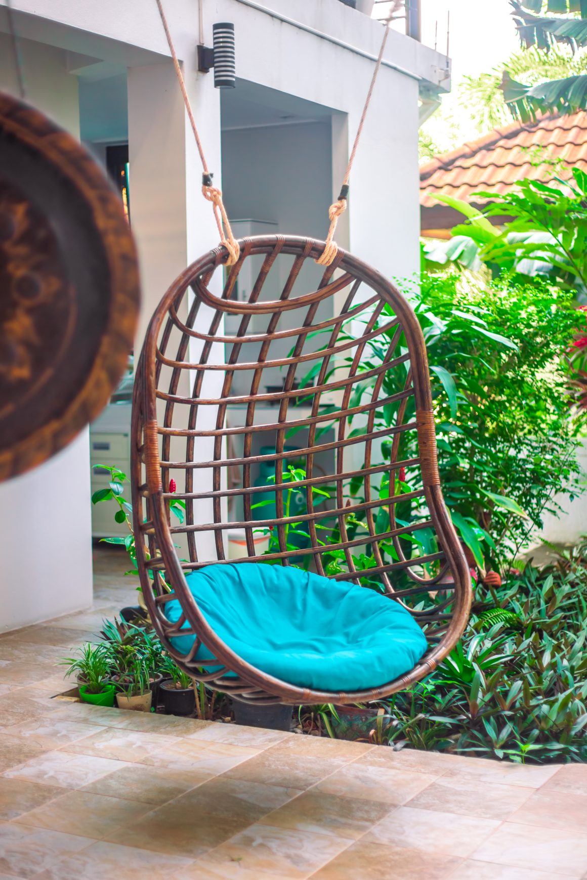 Terrasse avec fauteuil suspendu en osier, coussin bleu et végétation luxuriante