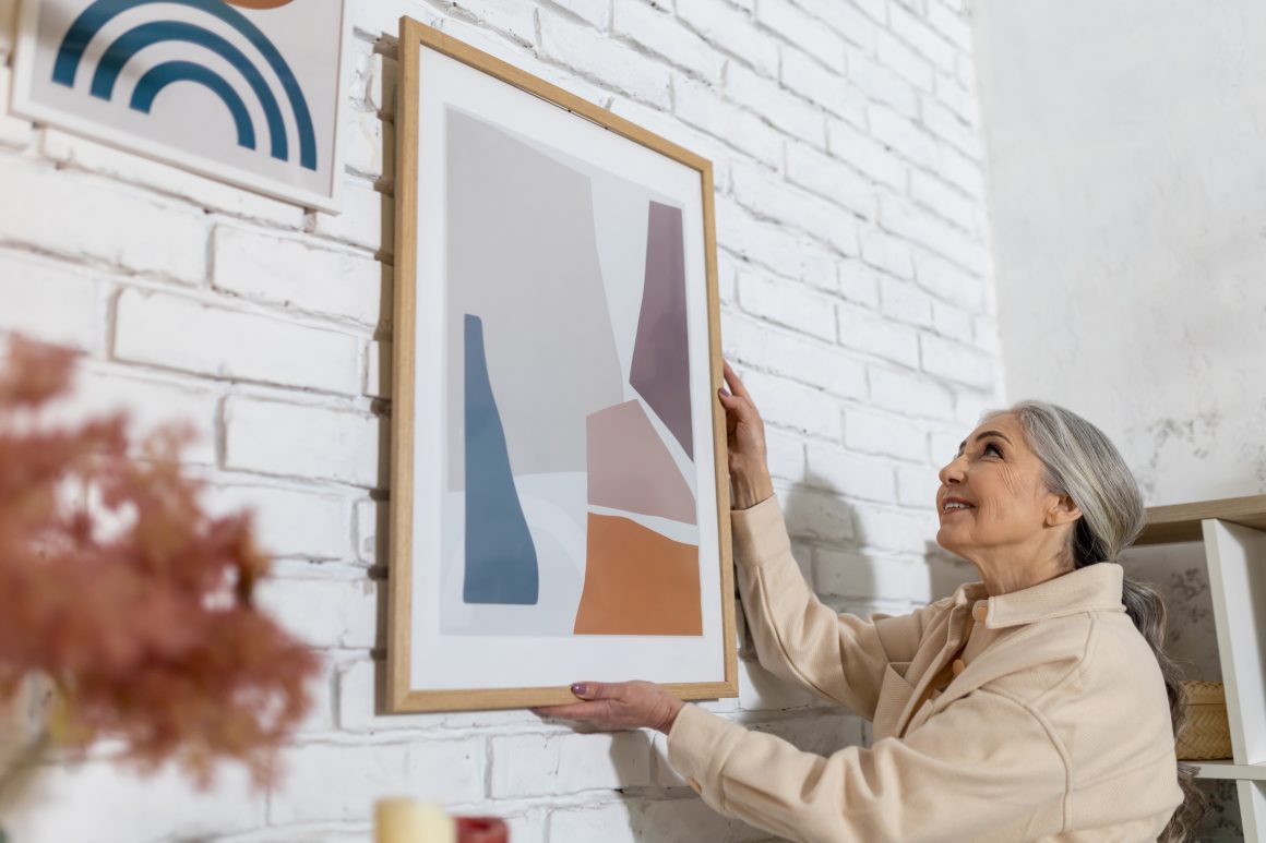 Une femme accroche une œuvre d'art abstraite encadrée sur un mur blanc en briques.
