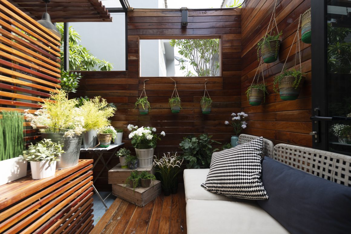 Petite terrasse cosy avec canapé, murs et sols en bois, nombreux pots de fleurs et plantes suspendues.