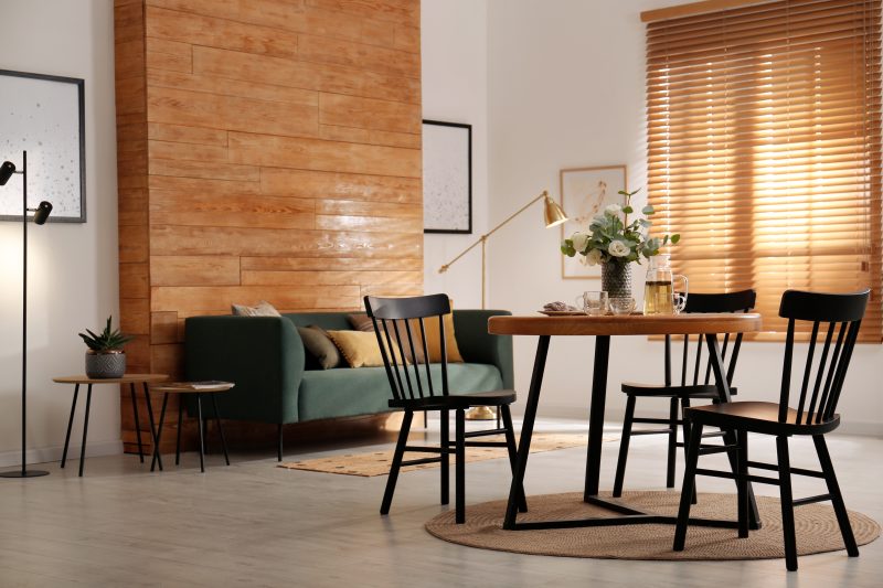 Aménager salon salle à manger 20m2 avec canapé vert, tables basses, table à manger avec chaises noires, tapis et stores en bois.