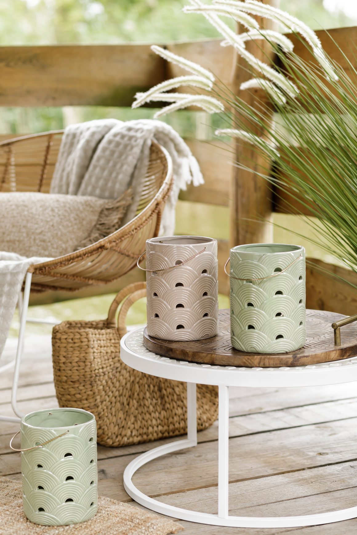 Un coin jardin cosy avec lanternes céramiques, table basse blanche, fauteuil rotin, coussins et plantes.