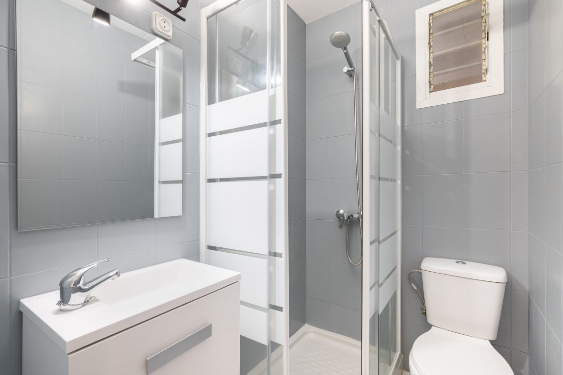 Salle de bain 4m2 avec douche cabine vitrée, lavabo, miroir, toilettes, carrelage gris et fenêtre d’aération.