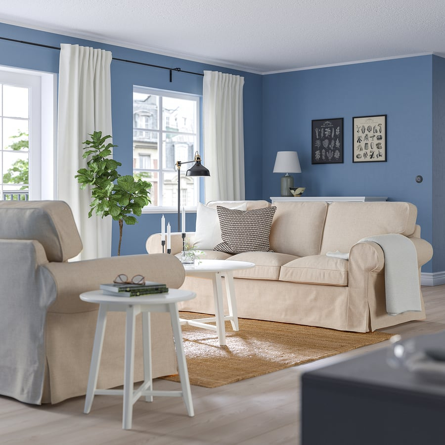Salon chaleureux avec murs bleus, canapé beige, table blanche, tapis naturel, plante verte et rideaux blancs.