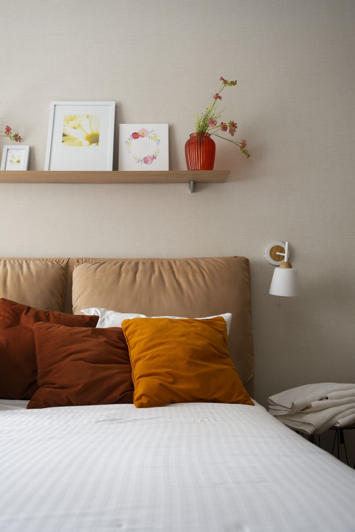 Lit beige avec coussins ocres et rouille et une étagère murale avec cadres et vase fleuri.