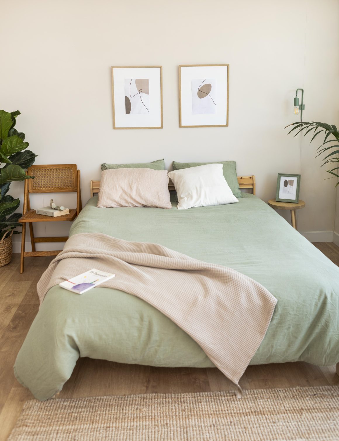 Petite chambre minimaliste avec lit vert sauge, plantes, chaises en bois et cadres muraux abstraits.