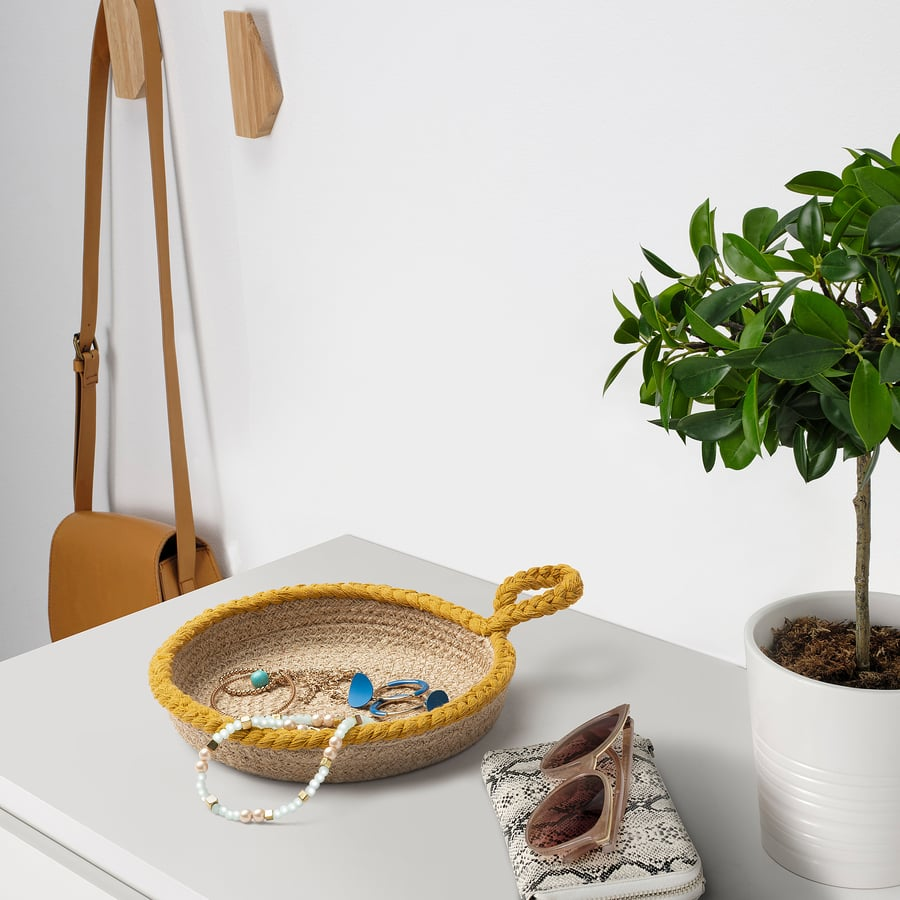 Panier tressé jaune contenant bijoux, pochette imprimée avec lunettes, plante verte et sac marron.