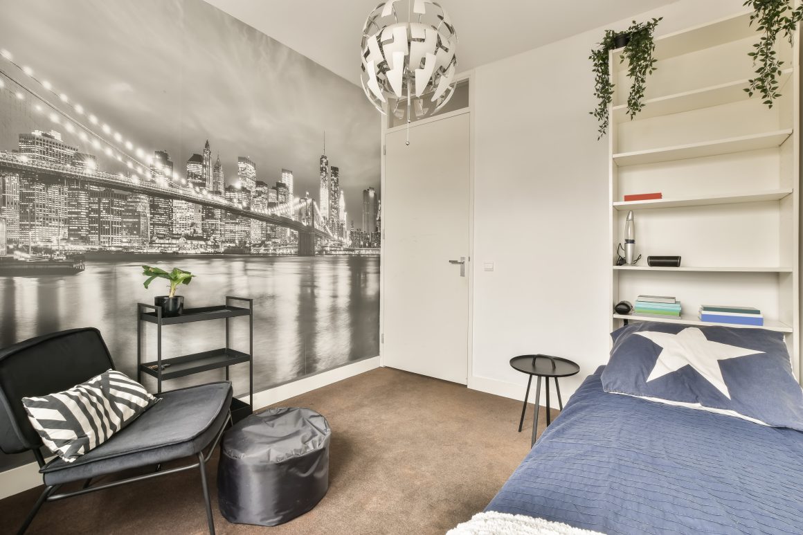 Chambre moderne avec un lit, fauteuil, mur panoramique skyline, étagères, table et plantes suspendues