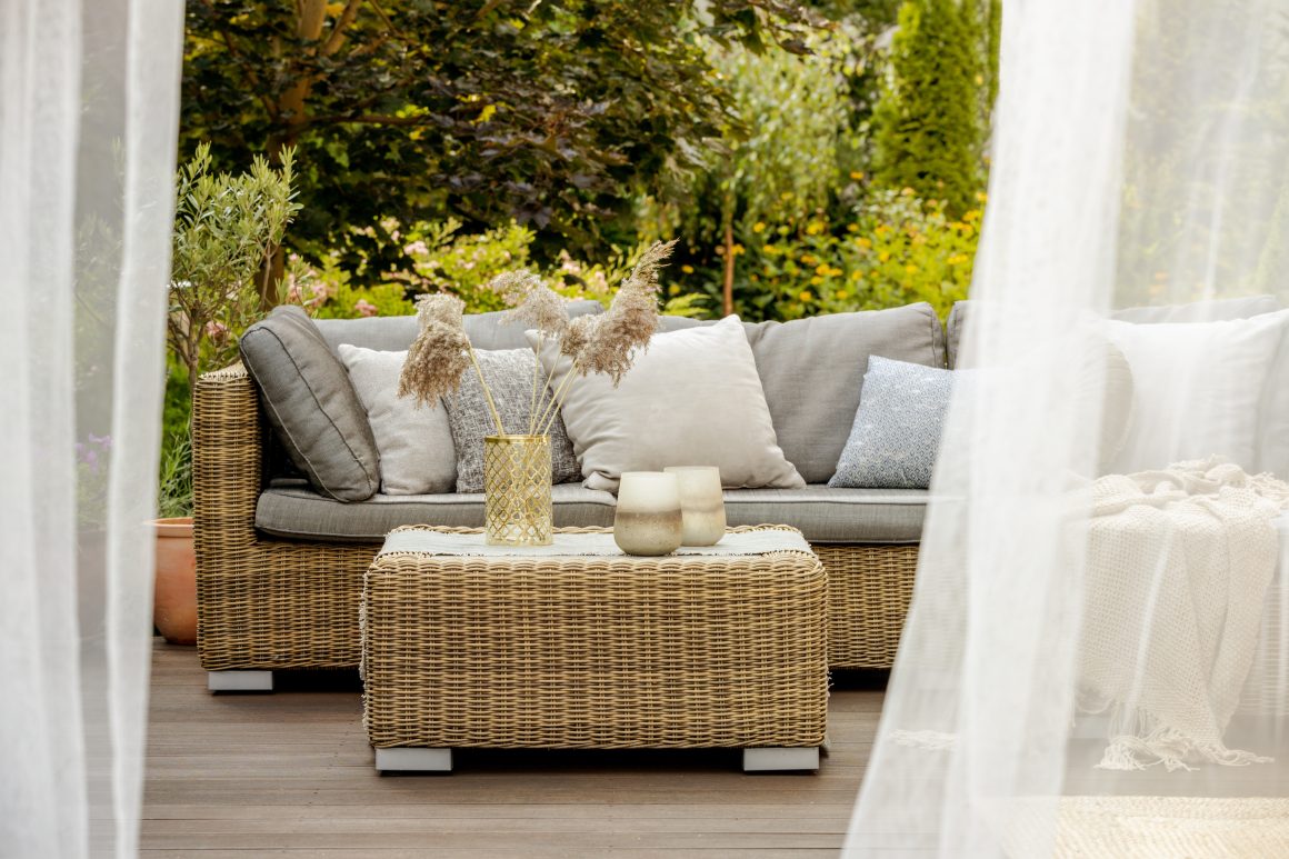 Salon de jardin extérieur avec canapé et table en osier, coussins gris, décor naturel et rideaux légers.