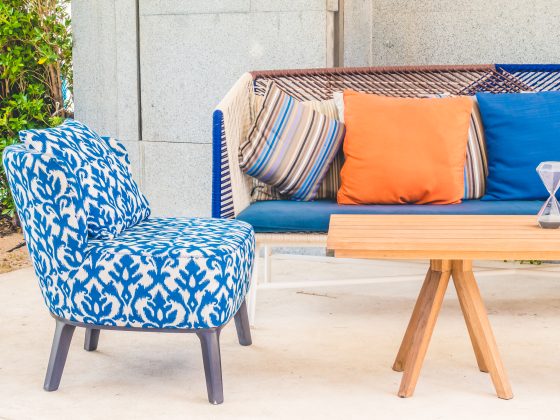 Chaise bleue à motifs, canapé coloré avec coussins orange, bleu, rayé, table en bois, sableuse et décor extérieur lumineux.