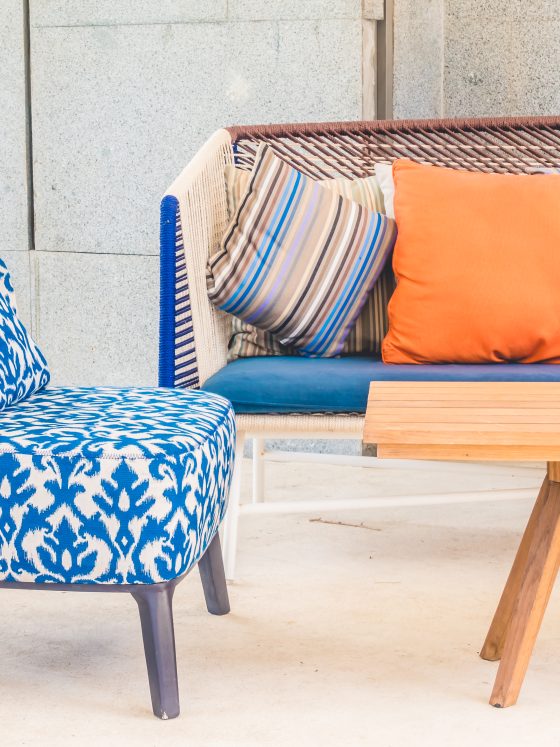 Chaise bleue à motifs, canapé coloré avec coussins orange, bleu, rayé, table en bois, sableuse et décor extérieur lumineux.