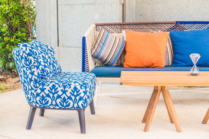Chaise bleue à motifs, canapé coloré avec coussins orange, bleu, rayé, table en bois, sableuse et décor extérieur lumineux.