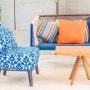 Chaise bleue à motifs, canapé coloré avec coussins orange, bleu, rayé, table en bois, sableuse et décor extérieur lumineux.