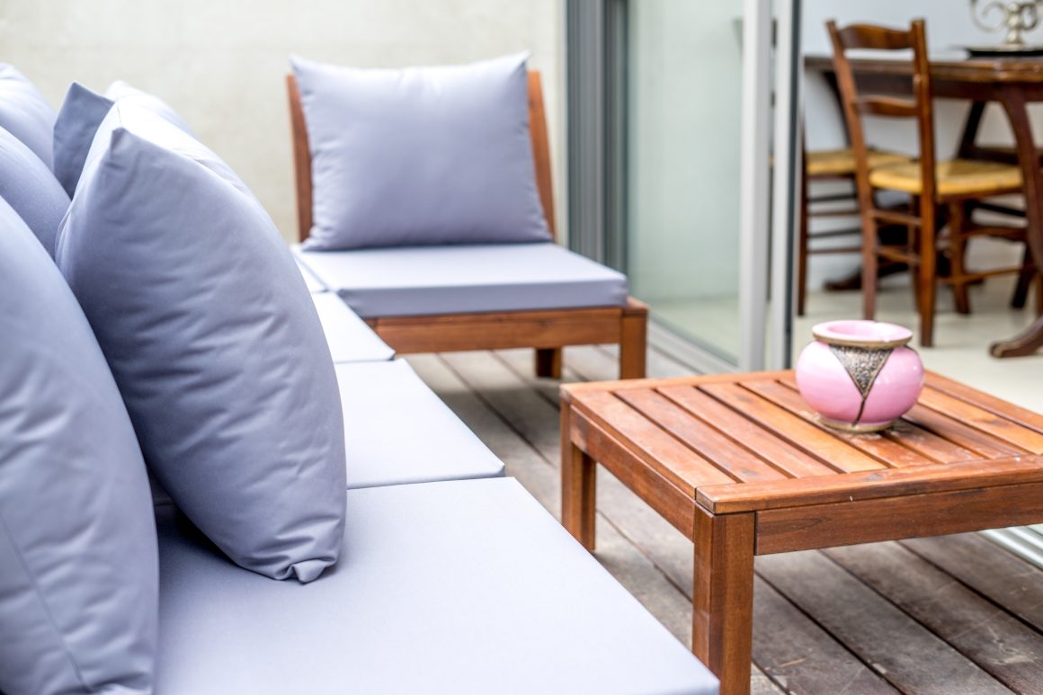 Un salon extérieur élégant avec canapés gris, coussins moelleux, table en bois et vase rose décoratif sur terrasse lumineuse.
