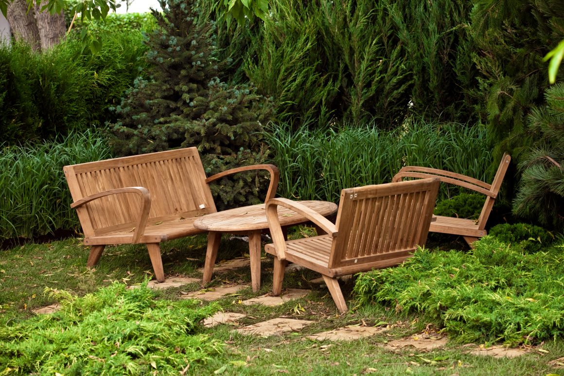 Un coin jardin paisible avec mobilier en bois : deux bancs, une chaise, une table ronde, entourés de verdure luxuriante.