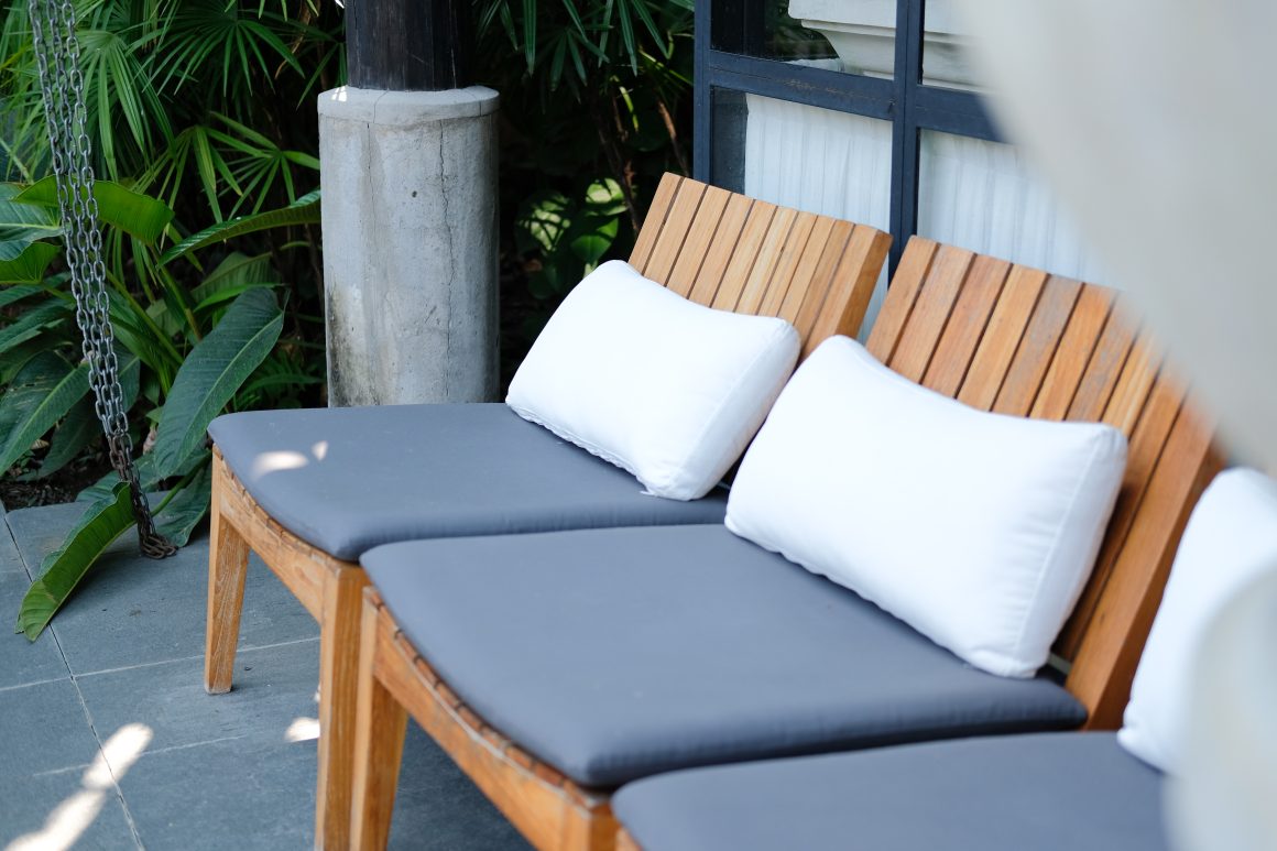 Chaise longue en bois avec coussins gris et oreillers blancs placée sur terrasse carrelée entourée de plantes vertes et béton
