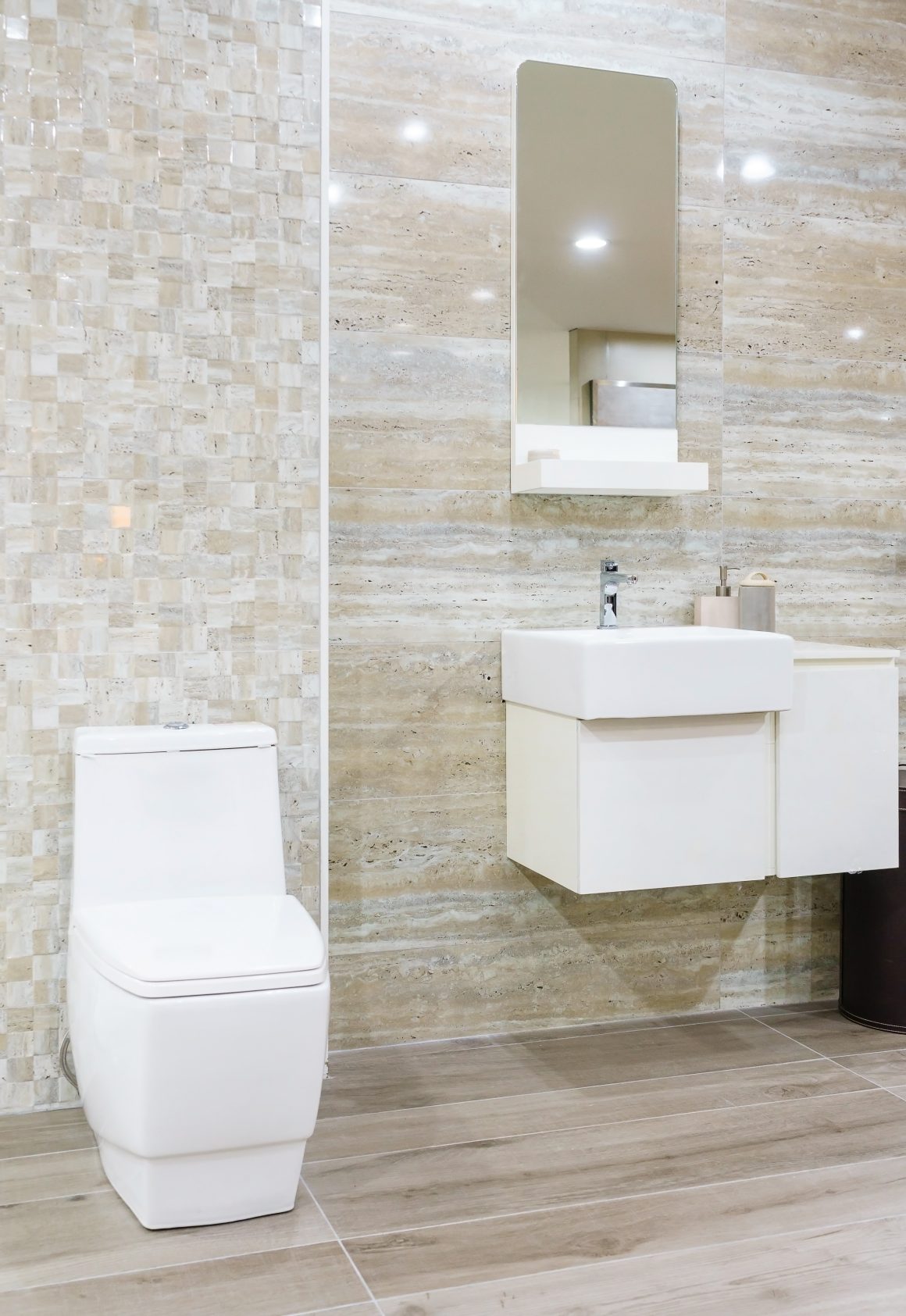 Salle de bain moderne avec toilettes, lavabo suspendu, miroir, mur en marbre beige et sol en bois clair.