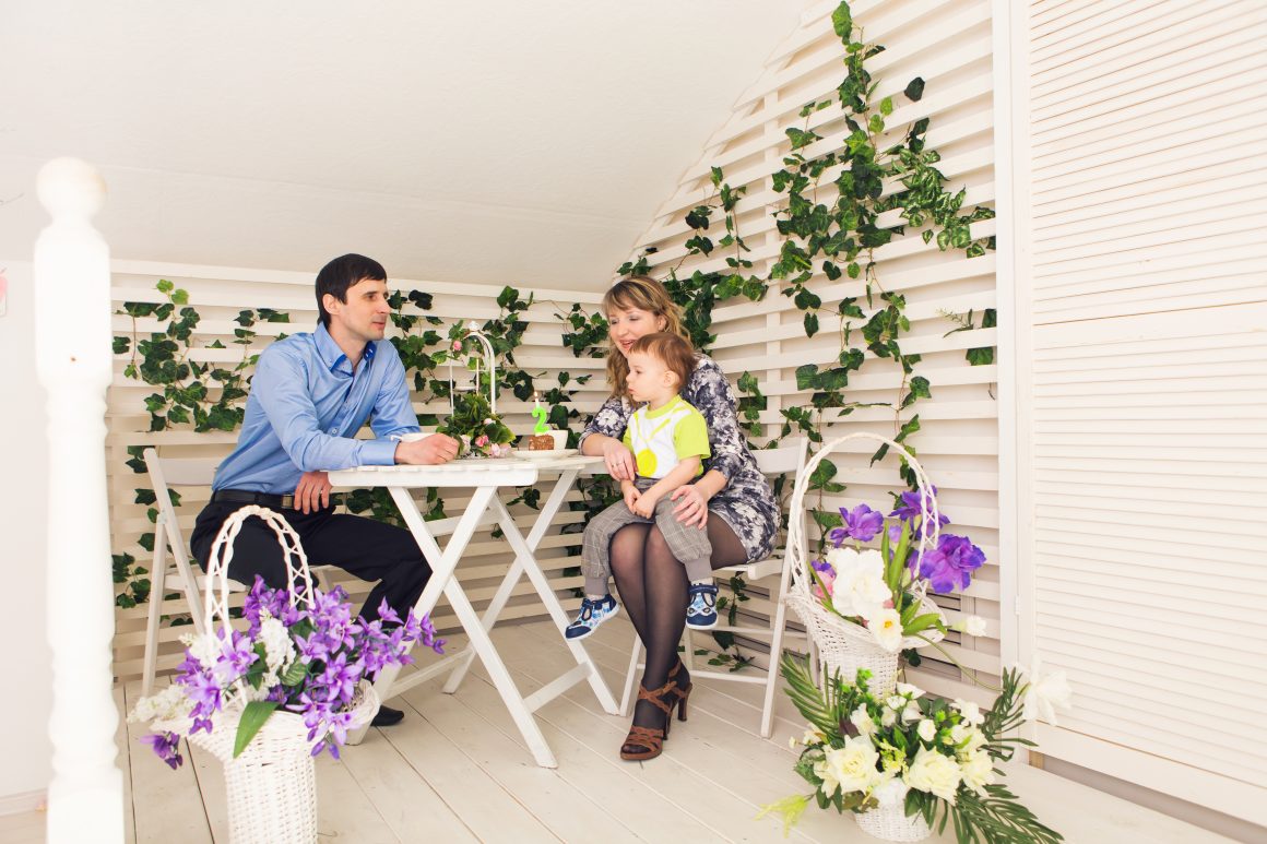Famille souriante sur terrasse lumineuse décorée de fleurs, plantes grimpantes et mobilier blanc