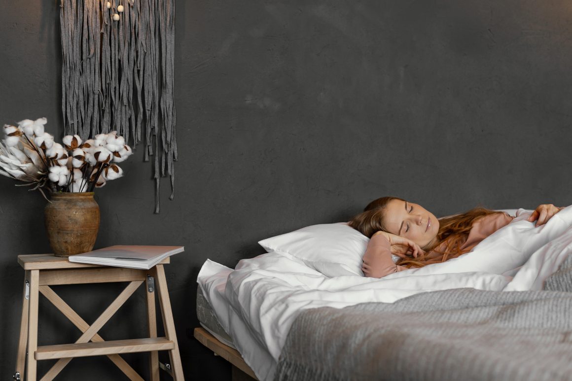 Jeune femme endormie dans une chambre apaisante incluant lit blanc, décor gris, table en bois et vase de coton.