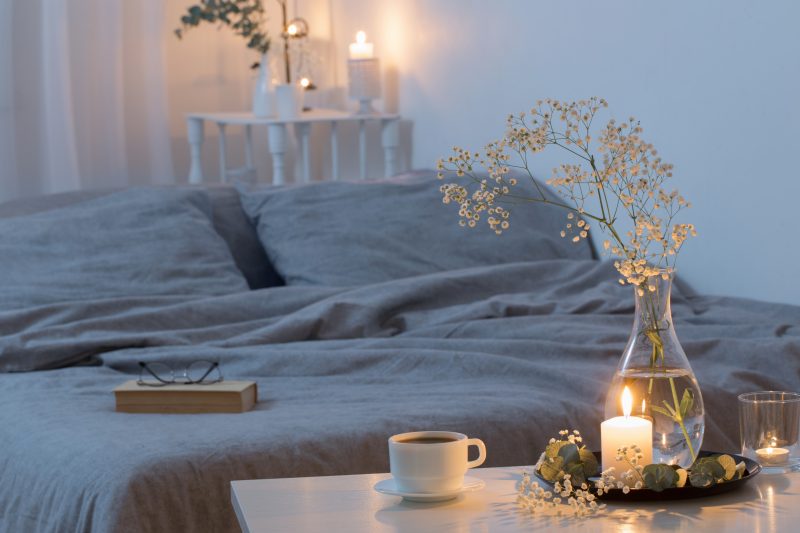 Chambre incluant lit gris, bougie allumée, vase avec fleurs : atmosphère apaisante et relaxante