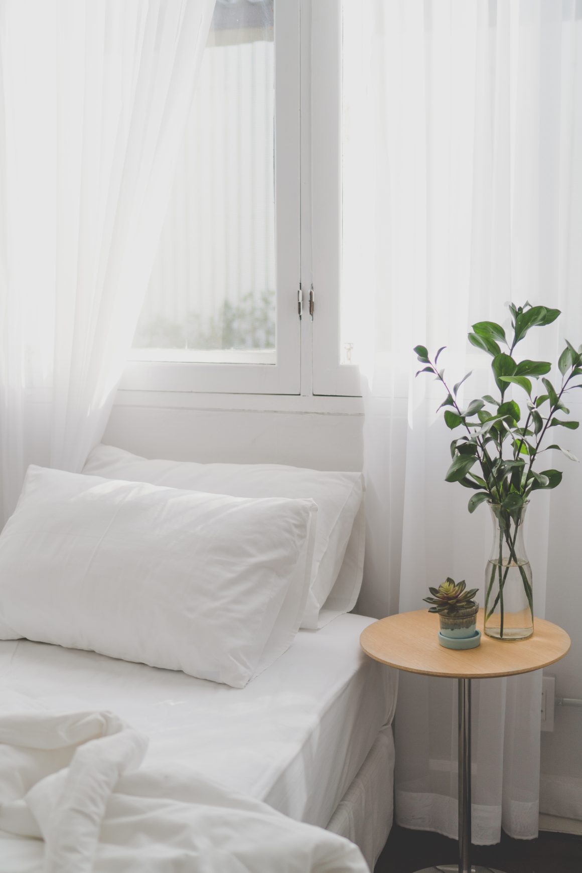 Chambre lumineuse incluant lit blanc, voilage, table ronde en bois et plante verte