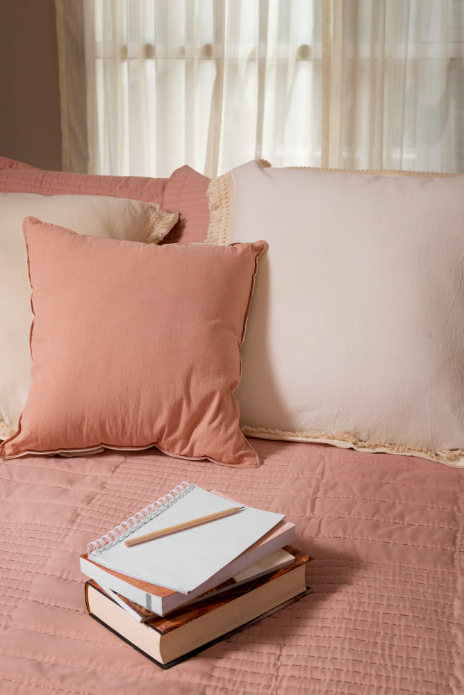 des coussins nude sur un lit confortable, avec deux cahiers dessus, devant une grande fenêtre
