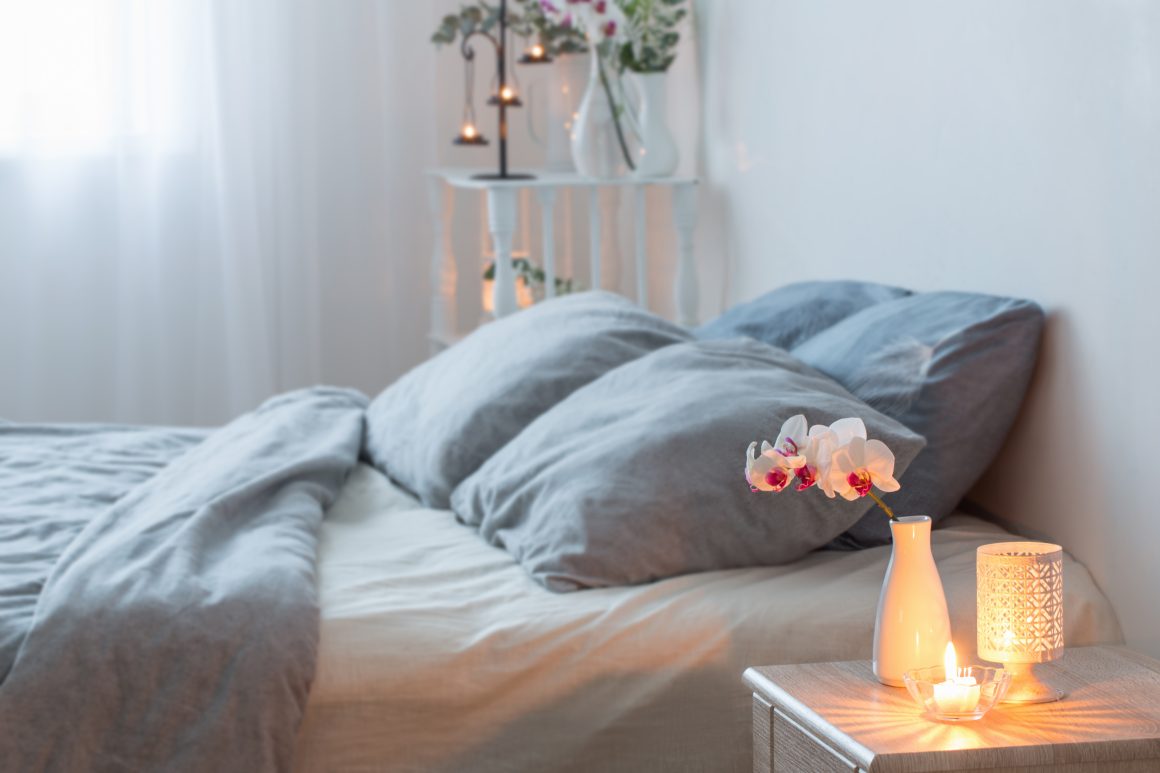 Chambre douce et lumineuse, lit gris, oreillers moelleux, vase avec orchidée et bougies allumées