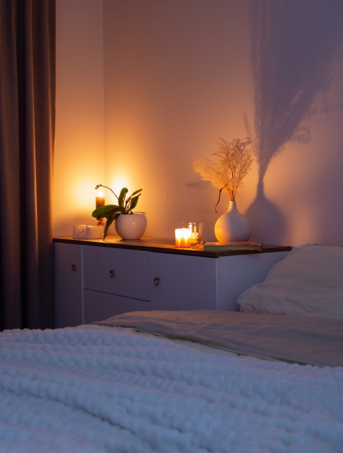 Une chambre chaleureuse éclairée par des bougies, avec lit, plantes et vase décoratif sur commode.