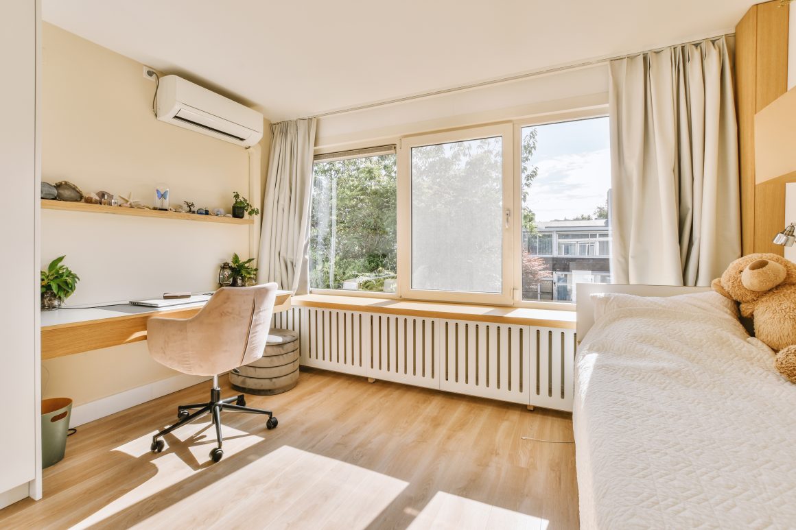 Chambre claire et cosy incluant bureau intégré, chaise rose, grande fenêtre, plantes, radiateur et rideaux