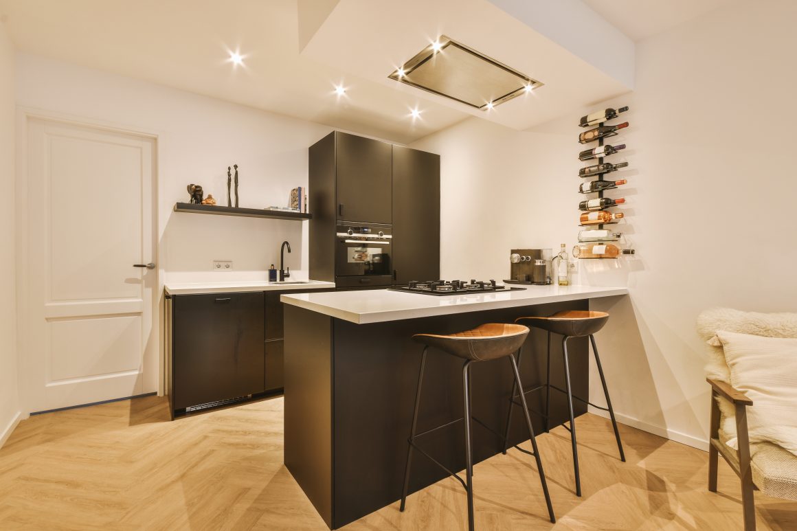 Cuisine moderne avec îlot central noir, tabourets en bois, rangement vertical pour vins et éclairage encastré