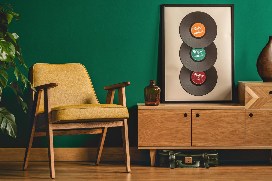 Coin salon rétro: fauteuil jaune, meuble en bois, vinyle mural, plante, bouteille, valise verte sous le meuble.