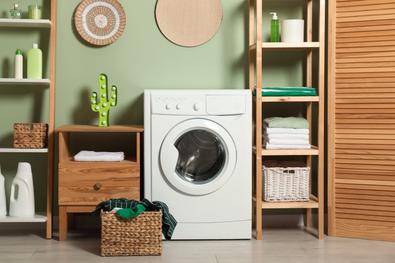 Aménagement garage en buanderie avec lave-linge blanc encadré d'étagères en bois, paniers tressés, serviettes et décor vert apaisant