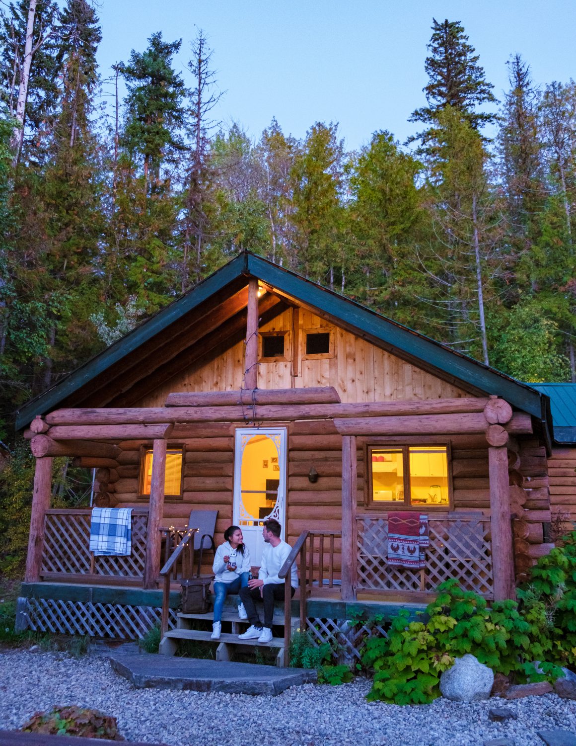 Chalet en rondins chaleureux, couple souriant sur la terrasse et forêt dense en arrière‑plan