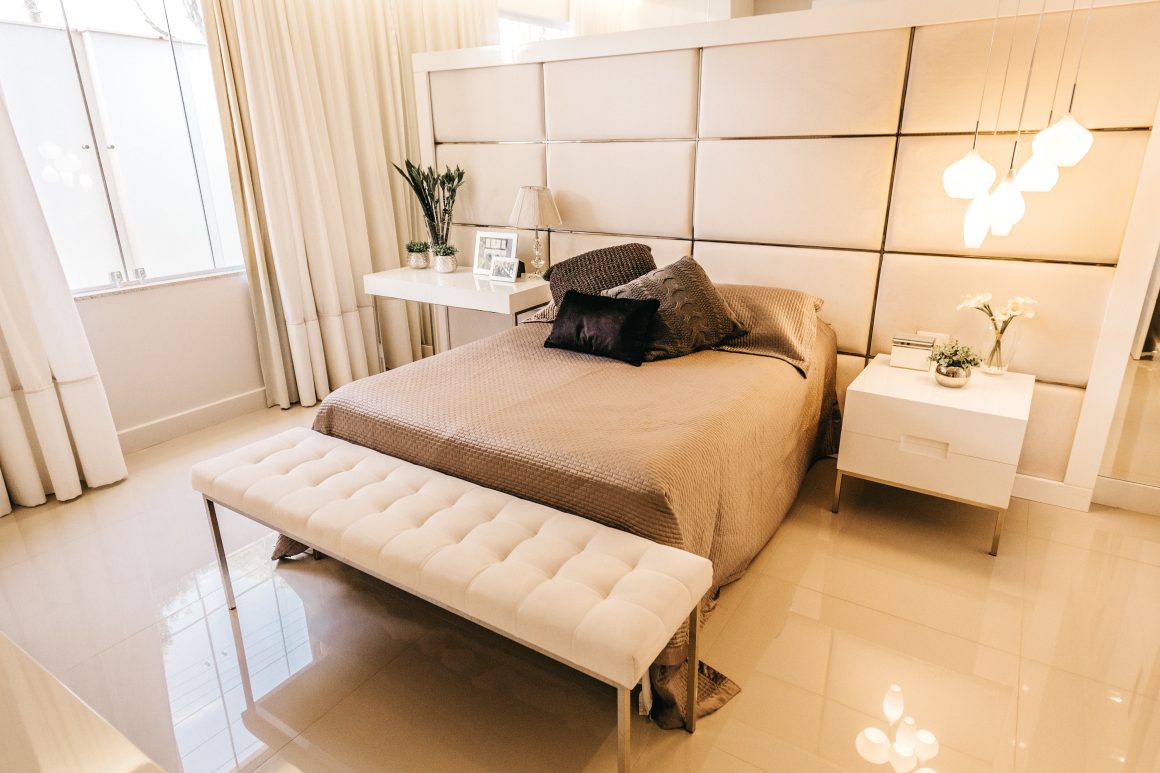 Chambre moderne aux tons neutres incluant lit avec banquette capitonnée, tables de chevet, luminaires et rideaux épais