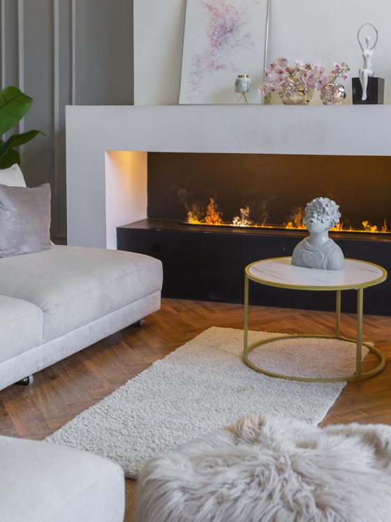 Salon cosy avec cheminée bio, canapés gris, table ronde dorée, plantes, tapis et objets décoratifs
