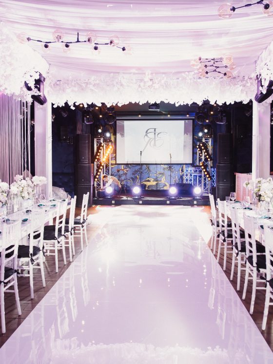 Décoration événement professionnel avec allée brillante, tables fleuries, chaises blanches et éclairage rose