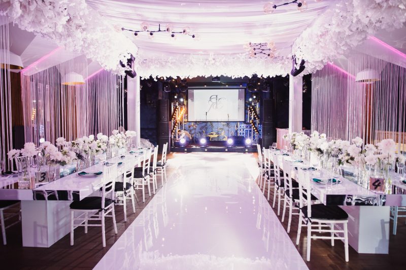 Décoration événement professionnel avec allée brillante, tables fleuries, chaises blanches et éclairage rose