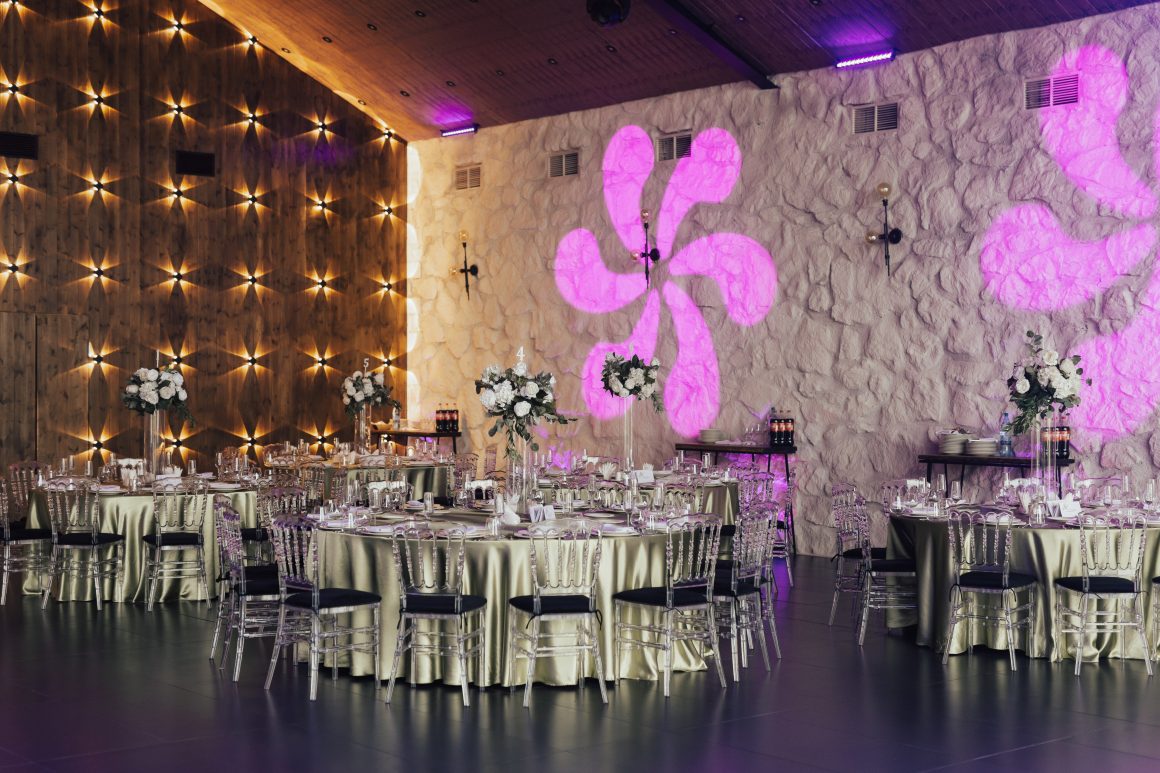 Salle de réception élégante avec tables drapées, chaises transparentes, centres floraux blancs, murs texturés et éclairage violet