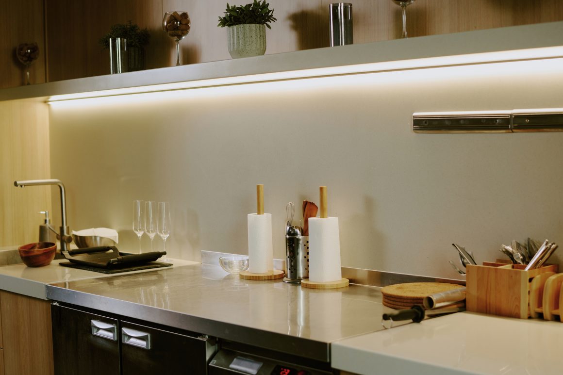 Plan de travail épuré en inox avec des ustensiles en bois, verres, plantes et éclairage doux sous placard