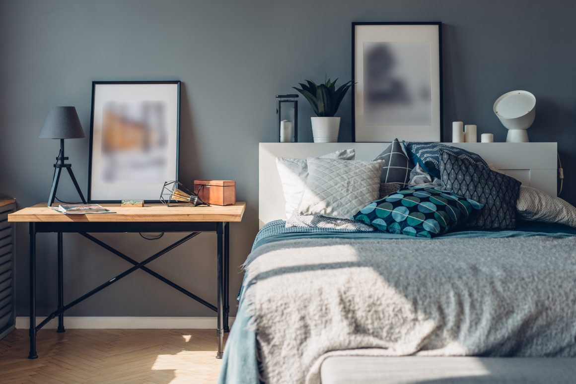 Chambre avec mur bleu gris, lit blanc avec coussins bleus et blancs, petit bureau en bois et objets décoratifs